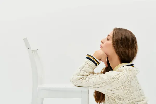 A pensive young woman rests her chin on her hand by a white chair in a calm setting. — Stock Photo