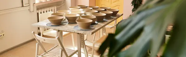 Table of unglazed pottery bowls for decoration and glazing, lively green plant. — Stock Photo