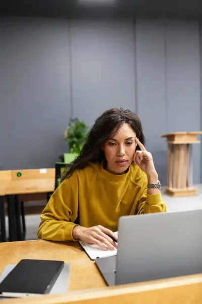 Elegant woman types thoughtfully on her laptop while seated in a stylish office environment. — Stock Photo