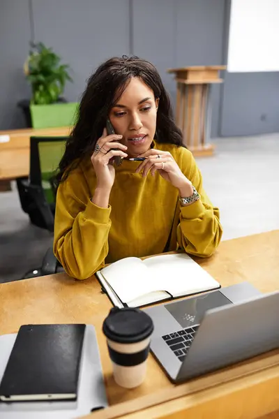 In un ambiente ufficio moderno, una bella donna lavora diligentemente alla sua scrivania, multitasking su una chiamata. — Foto stock