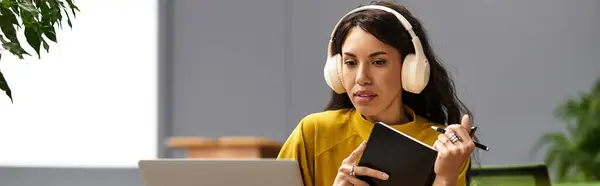 Hermosa mujer comprometida en tareas en su escritorio mientras usa auriculares y toma notas con atención. — Stock Photo