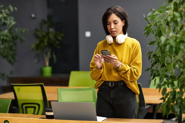 Elegant woman focuses on her smartphone while standing amidst a vibrant office environment. — Stock Photo