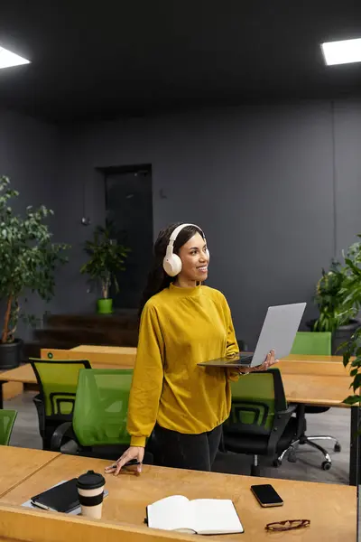 Beautiful woman wearing headphones stands in a stylish office space, engaged with her laptop. — Stock Photo