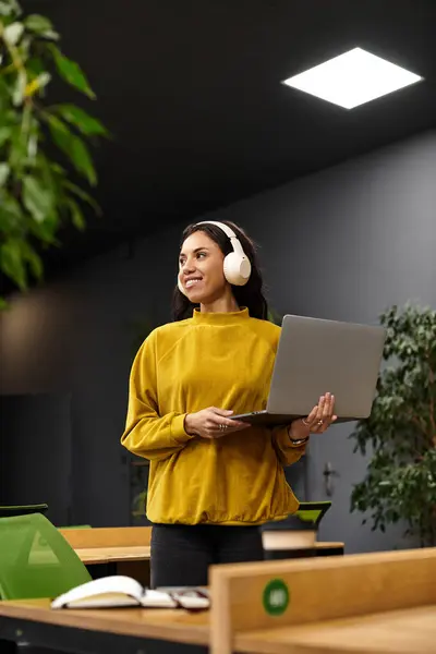 Young woman with stylish headphones smiles while using a laptop in a vibrant workspace. — Stock Photo