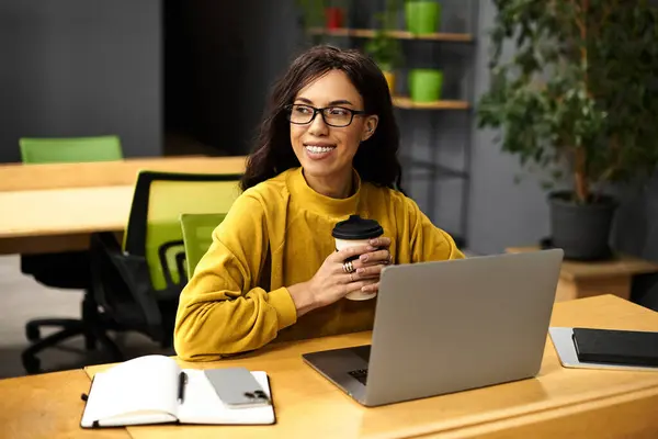 Young professional woman sips coffee while intently engaging with her laptop in a chic office. — Stock Photo
