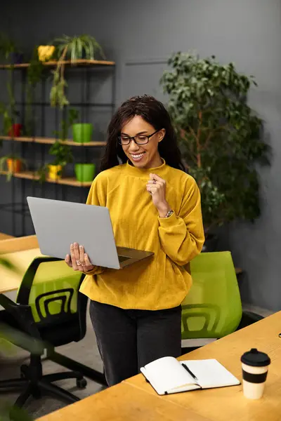 Uma mulher alegre em uma camisola brilhante equilibra seu laptop enquanto trabalhava em um escritório moderno. — Fotografia de Stock
