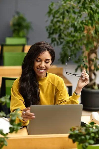 In a vibrant office space, a beautiful woman smiles while engaged with her laptop amidst plants. — Stock Photo