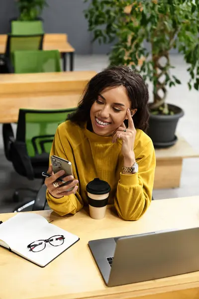 In a modern office, a young woman in a yellow sweater smiles as she uses her phone. — Stock Photo