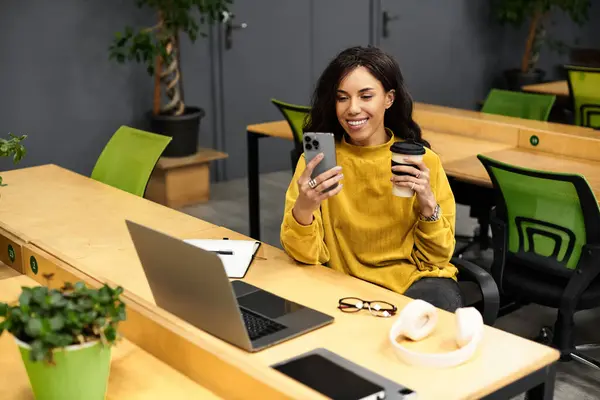 Una giovane ed elegante donna sorride al suo telefono sorseggiando un caffè in uno spazio di lavoro chic. — Foto stock
