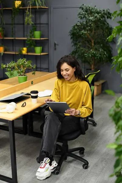 In a stylish office, an elegant woman smiles using a tablet, surrounded by greenery. — Stock Photo