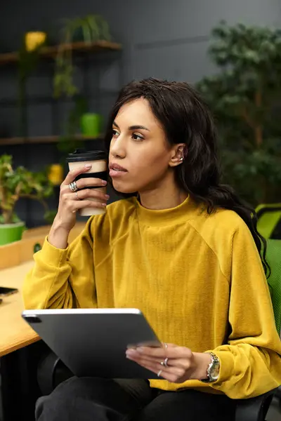 In un ufficio accogliente, una giovane donna gode il suo caffè mentre lavora intensamente sul suo tablet. — Stock Photo