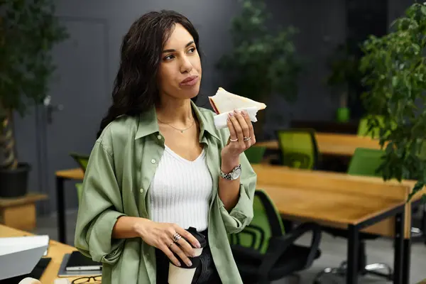 En una oficina moderna con plantas, una joven se detiene para disfrutar de su sándwich en el trabajo. — Stock Photo