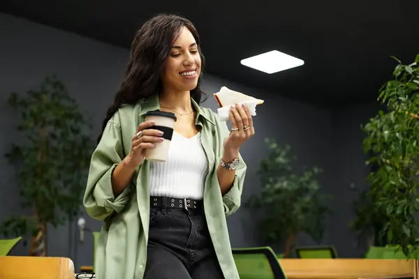Una giovane donna sorride mentre si gode un panino e un caffè durante la pausa ufficio. — Foto stock