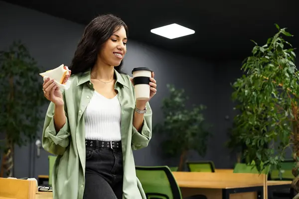 Eine junge, schöne Frau lächelt, während sie ein Sandwich und Kaffee in einem stilvollen Büro hält. — Stockfoto