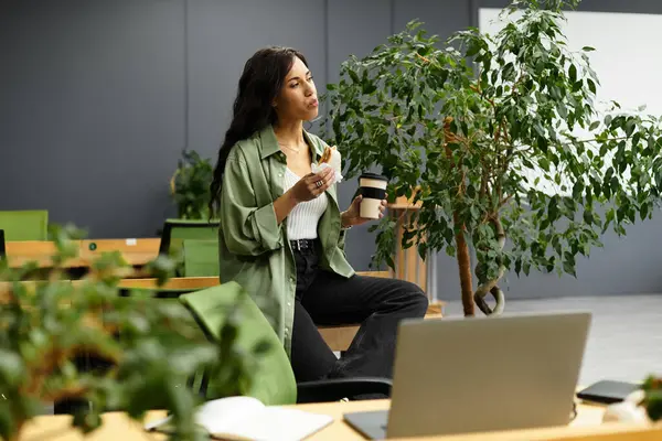 Beautiful woman takes a thoughtful moment with coffee and snack in contemporary office setting. — Stock Photo