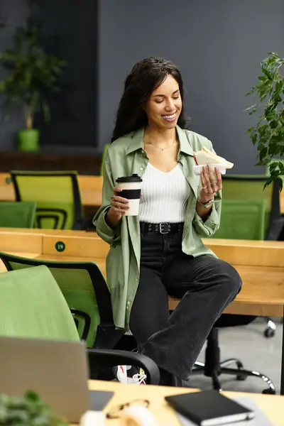 A joyful woman enjoys her coffee and snack in a green, modern office setting. — Stock Photo
