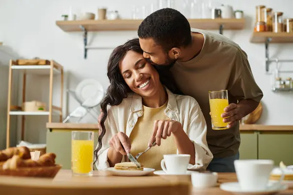 A man kisses a woman lovingly while she enjoys her breakfast at their cozy kitchen table. — Stock Photo