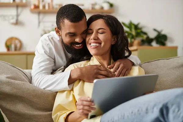 A joyful couple cuddles on a sofa, laughing and sharing affection while using a tablet. — Stock Photo