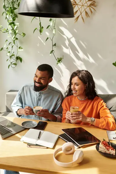 Una alegre pareja afroamericana disfruta del trabajo remoto juntos, bebiendo bebidas en un acogedor café. - foto de stock