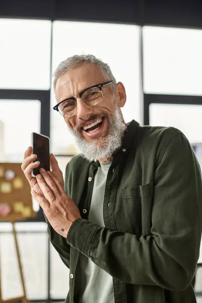Joyful man wearing glasses enthusiastically holds smartphone while chatting in bright office space. — Stock Photo