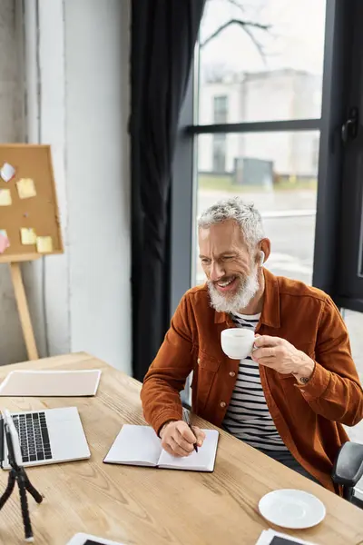 Un uomo felice si siede su una scrivania di legno, sorseggiando caffè e prendendo appunti in un ufficio luminoso. — Foto stock