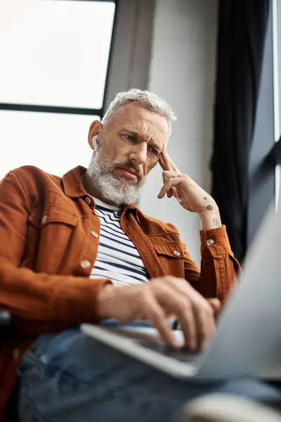 A man in casual attire types on his laptop while deep in thought in a well lit office. — Stock Photo