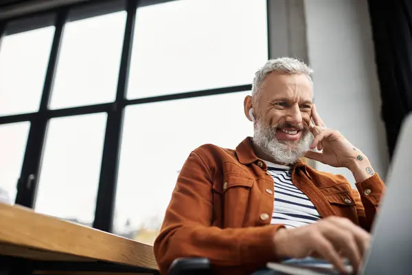 Un uomo con la barba che lavora felicemente al suo portatile in un ufficio illuminato dal sole, indossando auricolari senza fili. — Foto stock