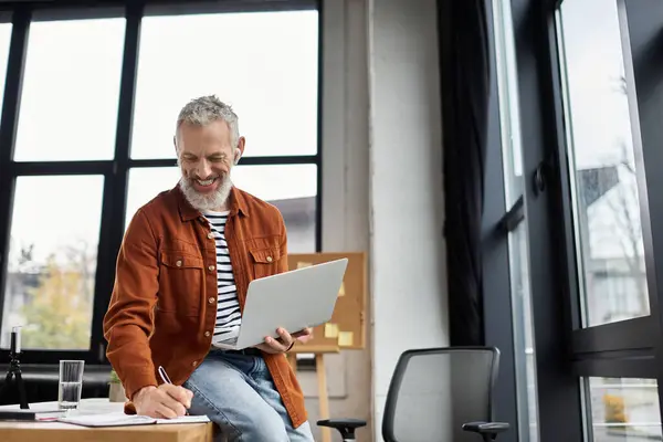 Un hombre sonriente con un portátil y un cuaderno disfruta de una productiva sesión de trabajo en una elegante oficina. - foto de stock