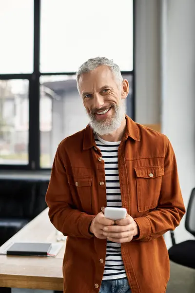 Un homme joyeux avec une barbe se tient dans un cadre de bureau élégant, engageant avec son smartphone. — Photo de stock