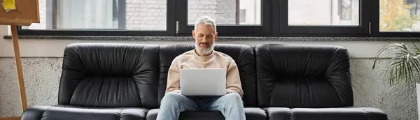Man sits comfortably on a leather couch, intently working on a laptop in a bright office space. — Stock Photo