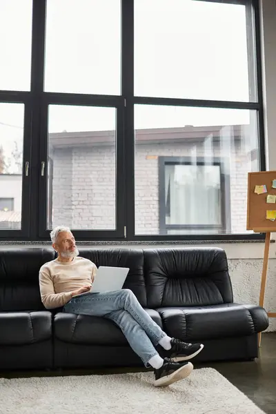 An older man sits comfortably on a black couch, focused on his laptop in a spacious office. — Stock Photo