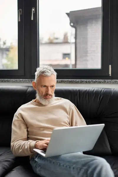 A man sits comfortably on a black couch, focused on his laptop in natural light. — Stock Photo