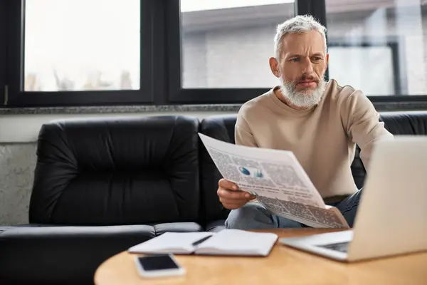 A focused individual sits on a couch, reading a newspaper while using a laptop for work. — Stock Photo