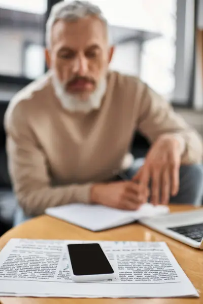 Person, die mit einem Smartphone, umgeben von Laptop und Notizen, sorgfältig Papiere überprüft. — Stockfoto