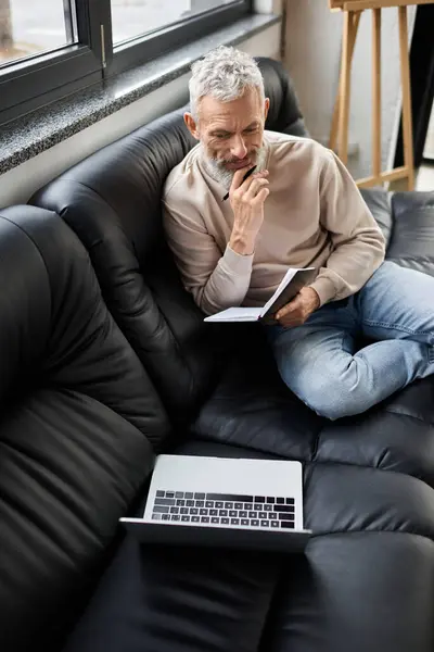 Focused individual sits on a couch, pondering ideas with a laptop and notebook at a workspace. — Stock Photo