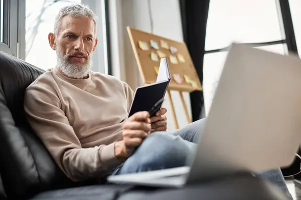 Man sits comfortably on a couch, reviewing notes while working on his laptop in a stylish office. — Stock Photo