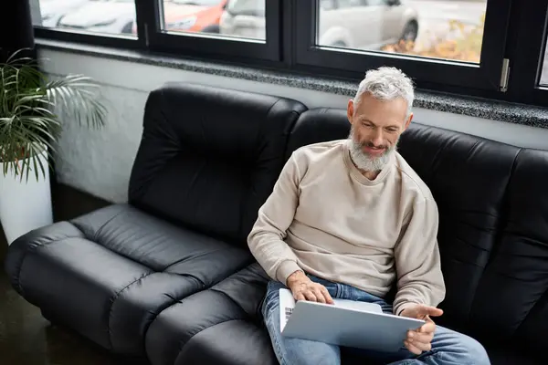 Um homem barbudo se senta em um sofá preto, focado em seu laptop em um escritório moderno. — Fotografia de Stock