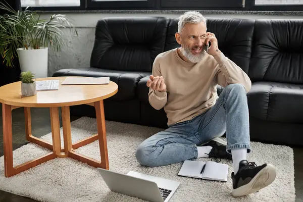 A mature man sits on the floor, chatting on the phone while using a laptop nearby. — Stock Photo