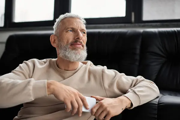 A man with a beard sits comfortably in an office, contemplating ideas while holding his phone. — Stock Photo