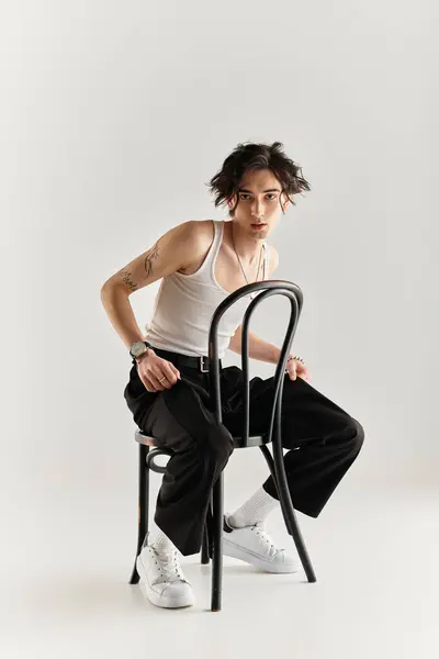 Um jovem magro senta-se em uma cadeira, exibindo um elegante top tanque branco e estilo moderno no estúdio. — Fotografia de Stock