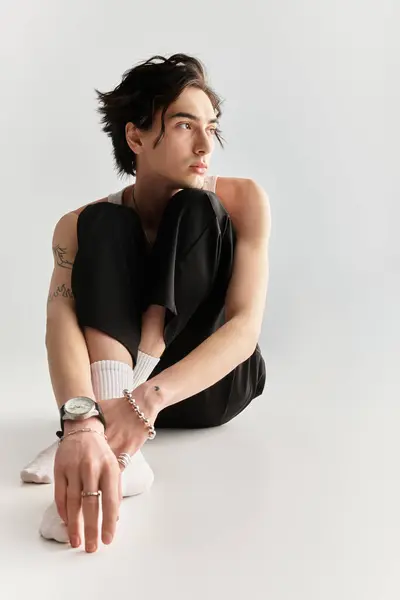 A slim young man sits on the studio floor, dressed in a stylish white tank top and black pants. — Stock Photo