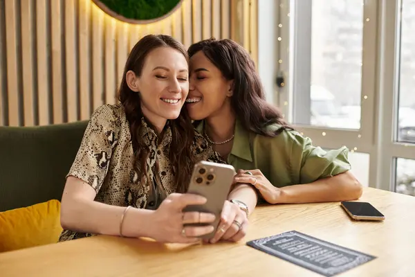 Two women share a joyful moment at a cafe, smiling and looking at a smartphone together. — Stock Photo