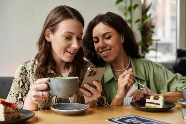 Due donne condividono un momento gioioso, gustando caffè e dessert mentre utilizzano uno smartphone. — Stock Photo