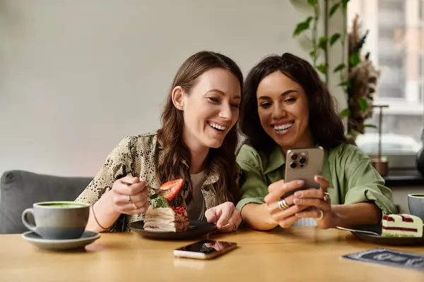 A joyful lesbian couple enjoys dessert at a cafe, laughing while engaging with a smartphone. — Stock Photo