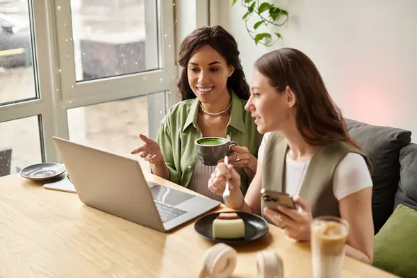 Two friends share laughter and delicious treats at a cafe while engaging with their devices. — Stock Photo
