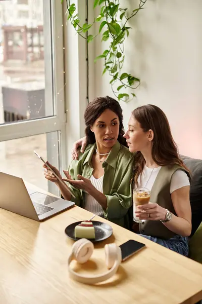 Two women share laughter and warm drinks at a cozy cafe, surrounded by greenery. — Stock Photo