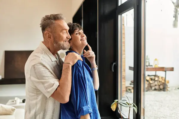 A husband and wife enjoy a warm moment, embracing each other by the large window. — Stock Photo