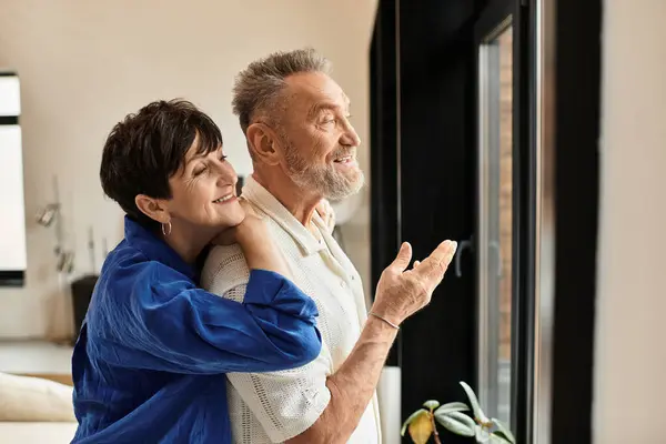 A joyful couple embraces each other while admiring the outdoors from their bright living space. — Stock Photo