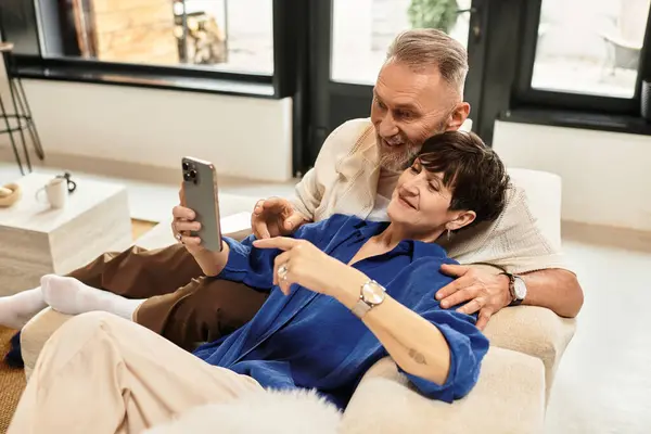 Middle aged couple shares laughter and joy while looking at a phone on the sofa. — Stock Photo