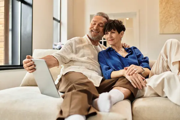 Un mari et sa femme sourient tout en partageant un moment confortable sur le canapé, en regardant une tablette ensemble. — Photo de stock